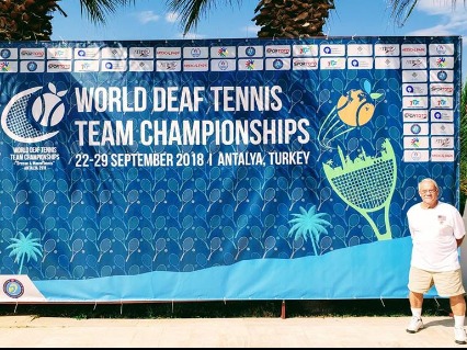 Howard Lee Gorrell poses for picture behind the huge banner - World Deaf Tennis Team Championships